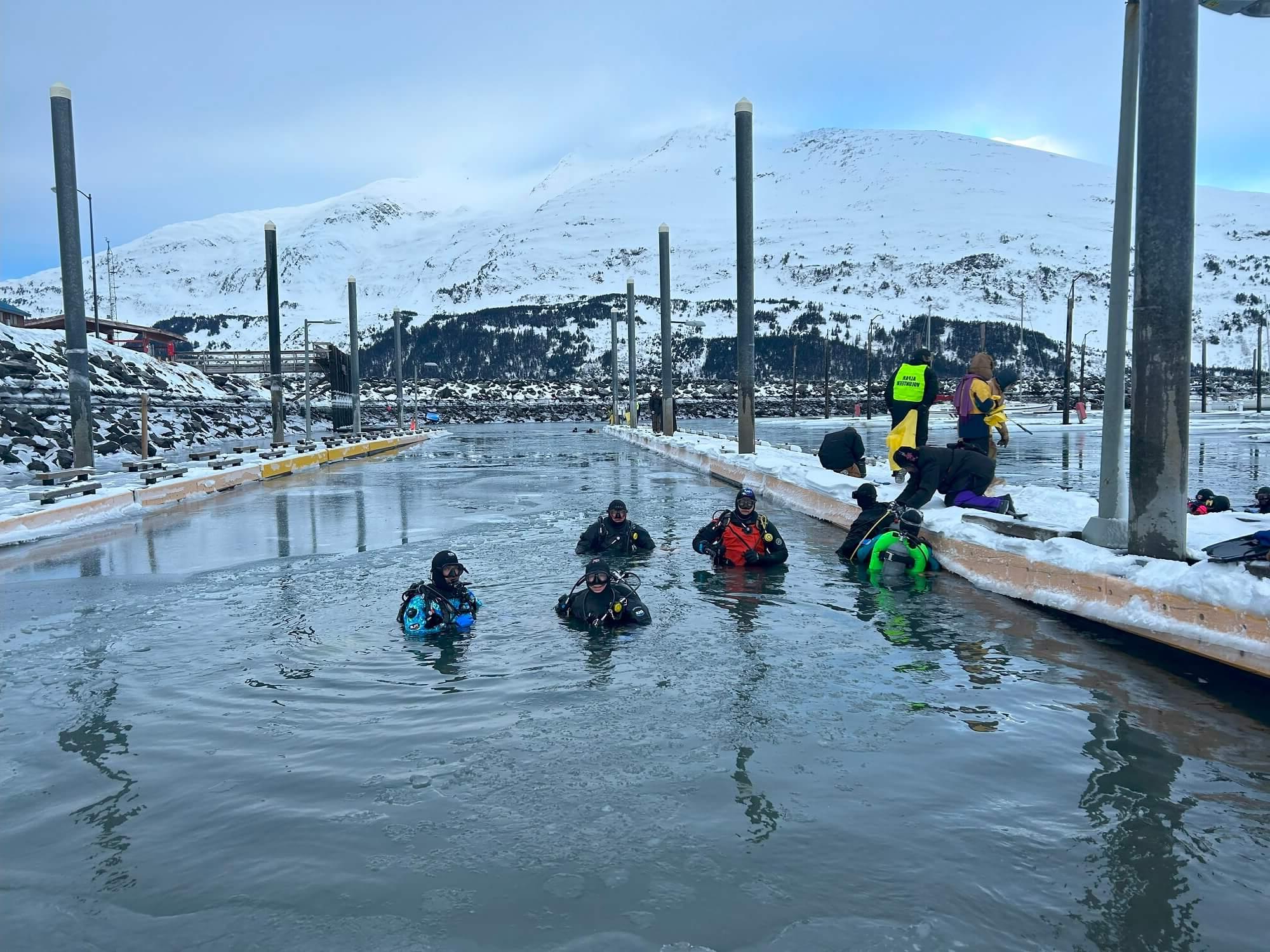 Divers stand in frigid Whittier Harbor waters with a snow capped mountain in the background.