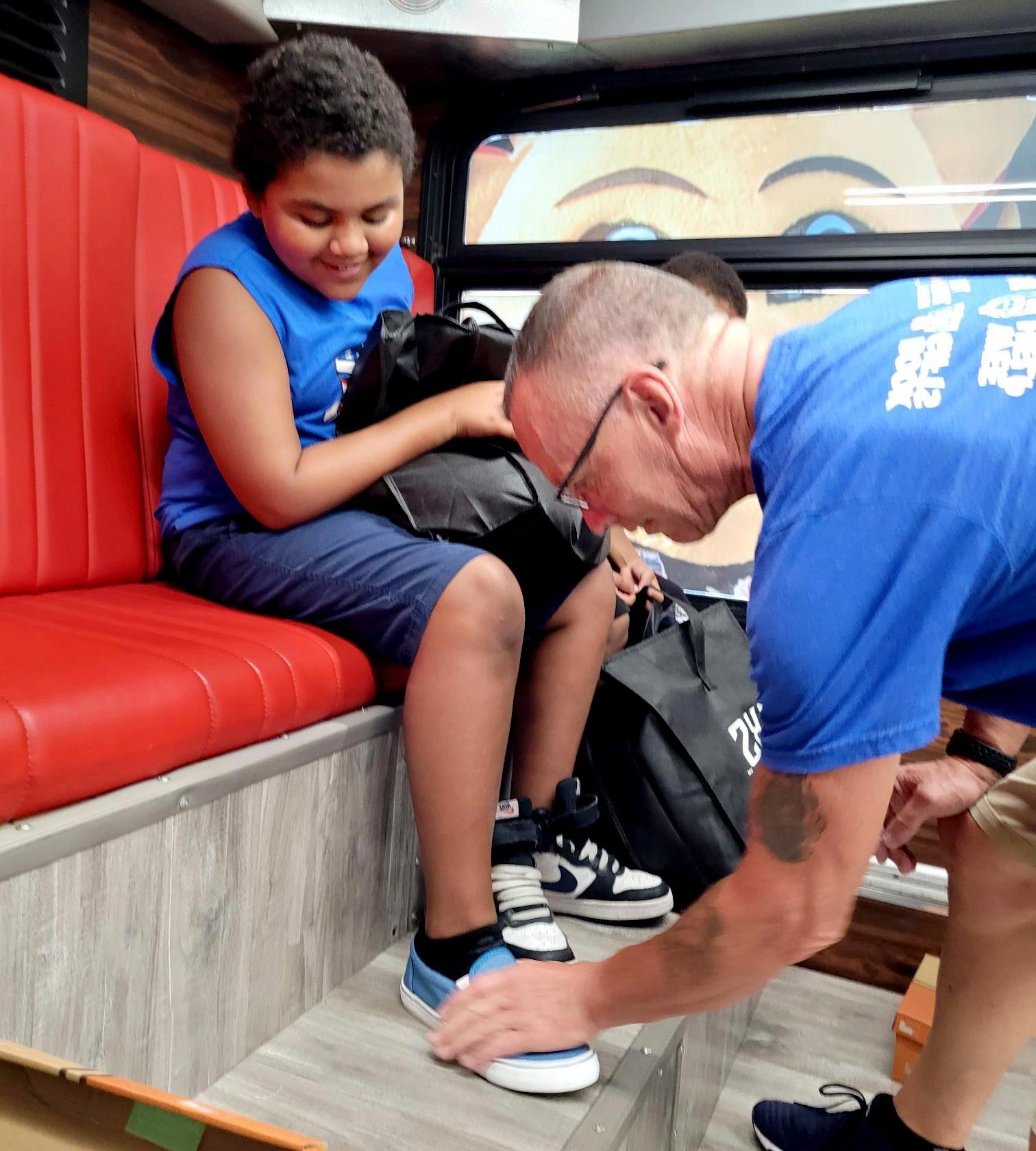 A young student seated on the red Delivering Dreams Bus bench tries on new shoes with the help of a volunteer.